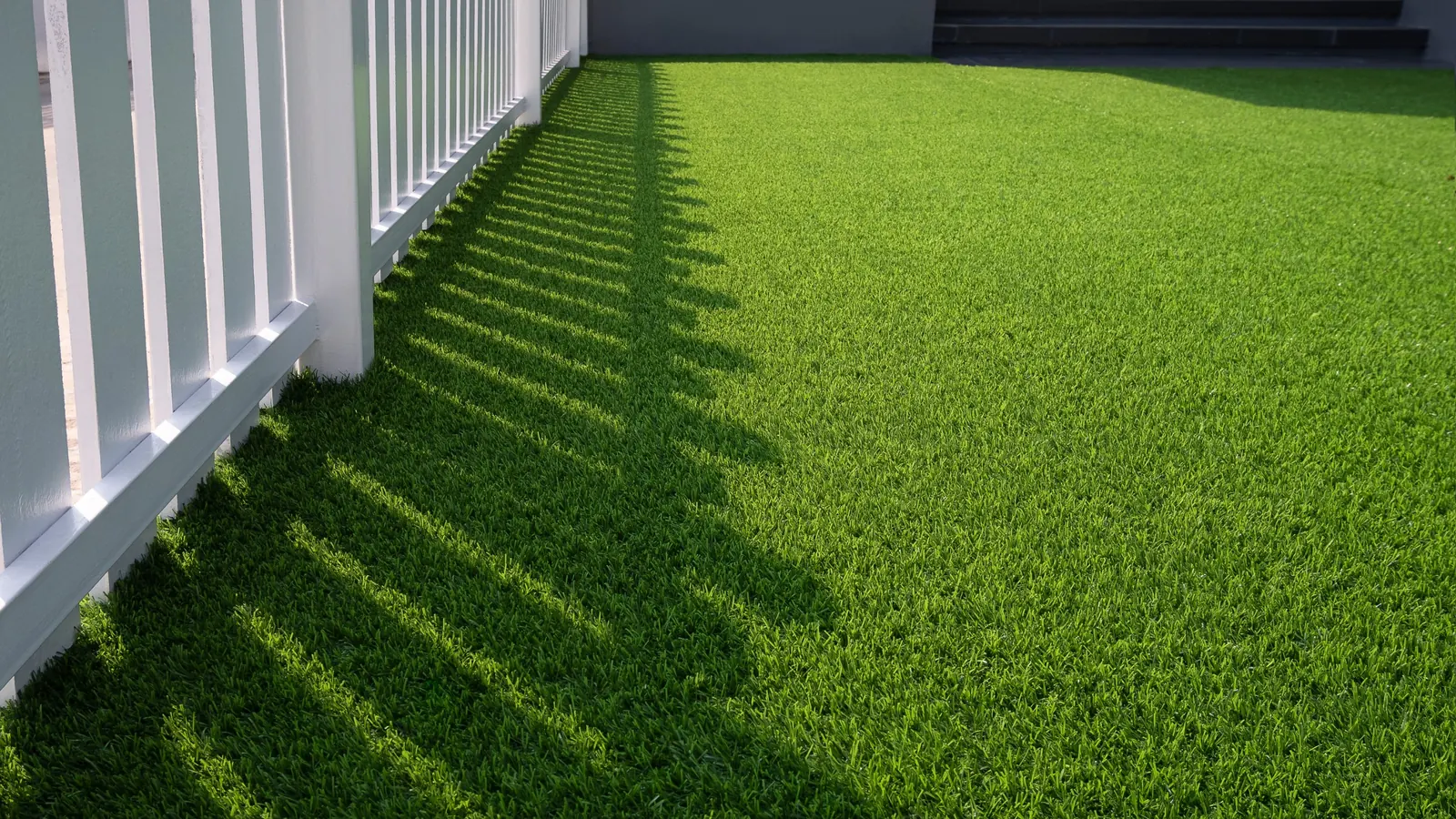 Artificial turf lawn with sunlight and fence shadows beside a white picket fence